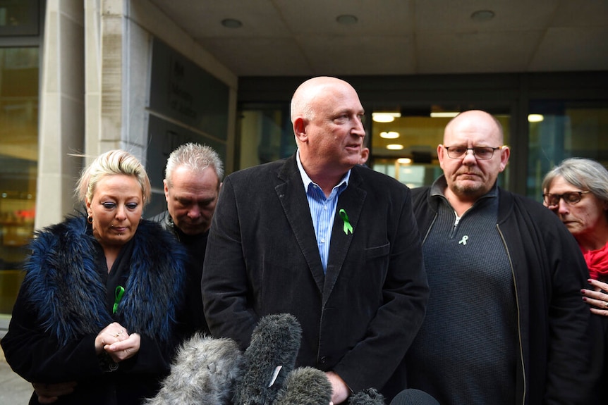 The Dunn family stand before a row of microphones while each wear a green lapel ribbon.