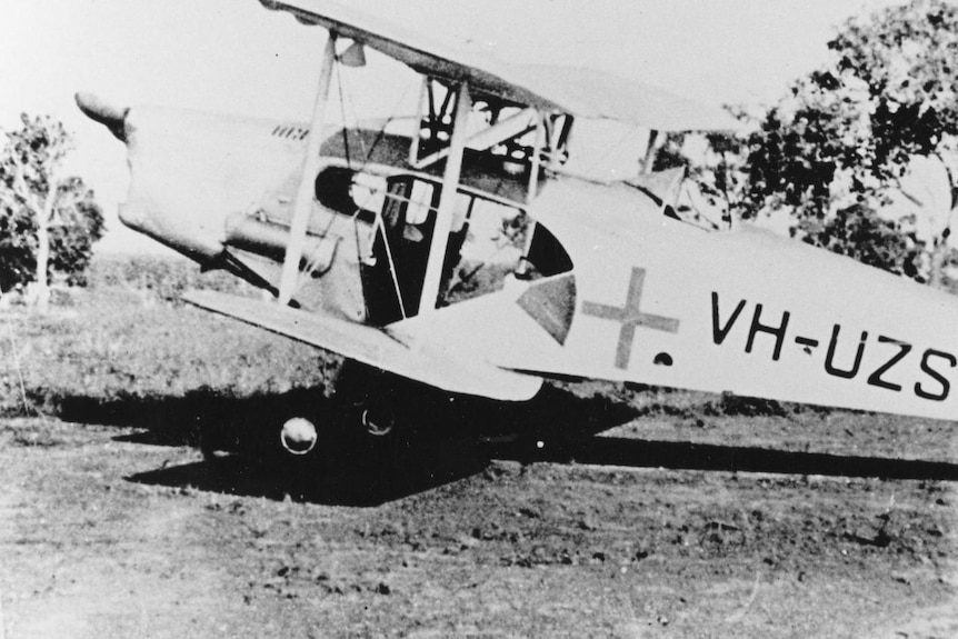 An old plane with a cross on it is landed in the bush.  The photo is old and in black and white. 