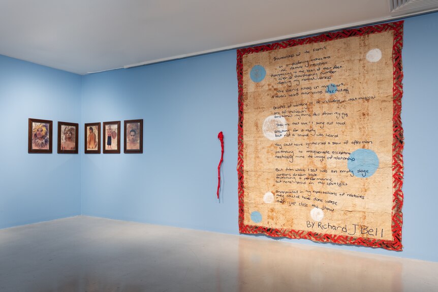 Large art work on Tongan bark cloth with red patterned trimmings hangs on blue wall besides  five smaller frames. 