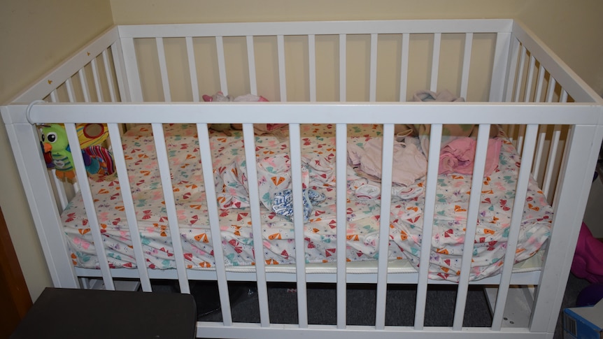 A blue and white cot inside a house with other objects and clothes on the carpet around it.