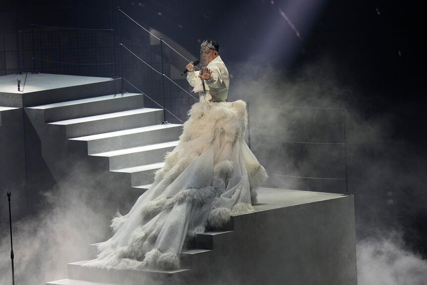 Australia's Sheldon Riley stands on a staircase wearing a crystal mask and a big white dress, singing his Eurovision song.