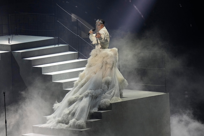 Sheldon Riley de Australia se para en una escalera con una máscara de cristal y un gran vestido blanco, cantando su canción de Eurovisión.
