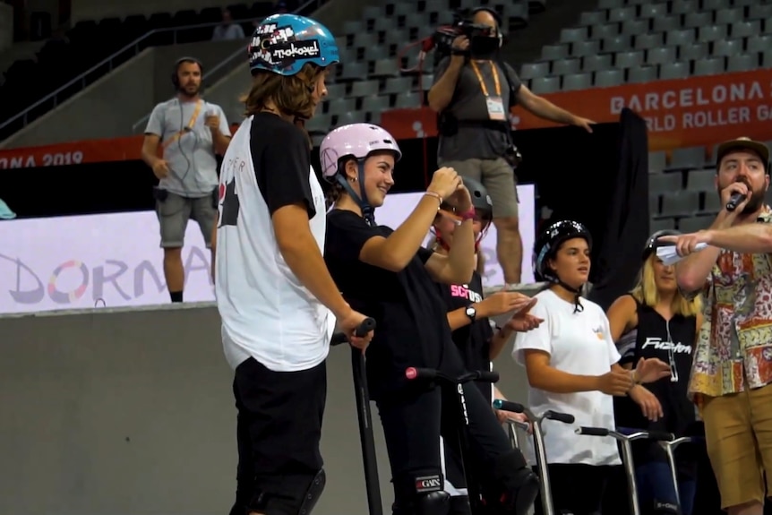 A girl wearing a pink helmet smiles while others holding scooters cheer her on.