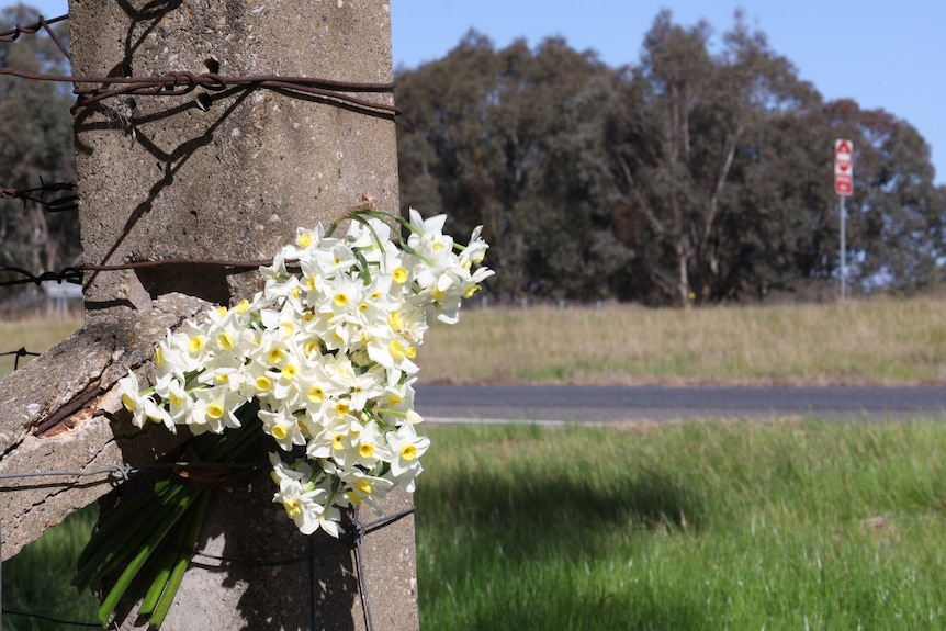 Flowers tied to a post, green grass and road in front, blue skies, trees.