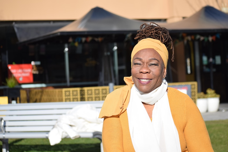 A woman smiling at the camera. She is wearing a yellow knitted top with a white shirt underneath. She wears her locs in a bun.