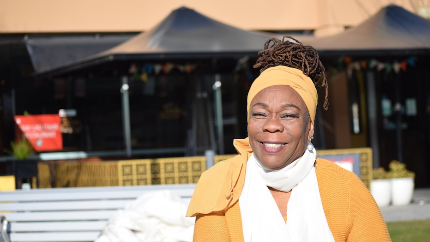 A woman smiling at the camera. She is wearing a yellow knitted top with a white shirt underneath. She wears her locks in a bun.