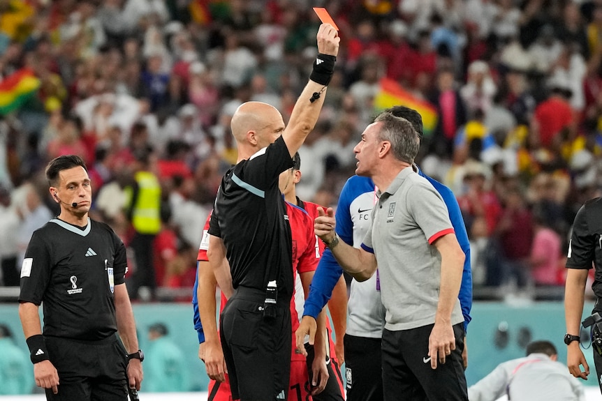 A referee holds a red card up towards South korea maneger Paulo Bento