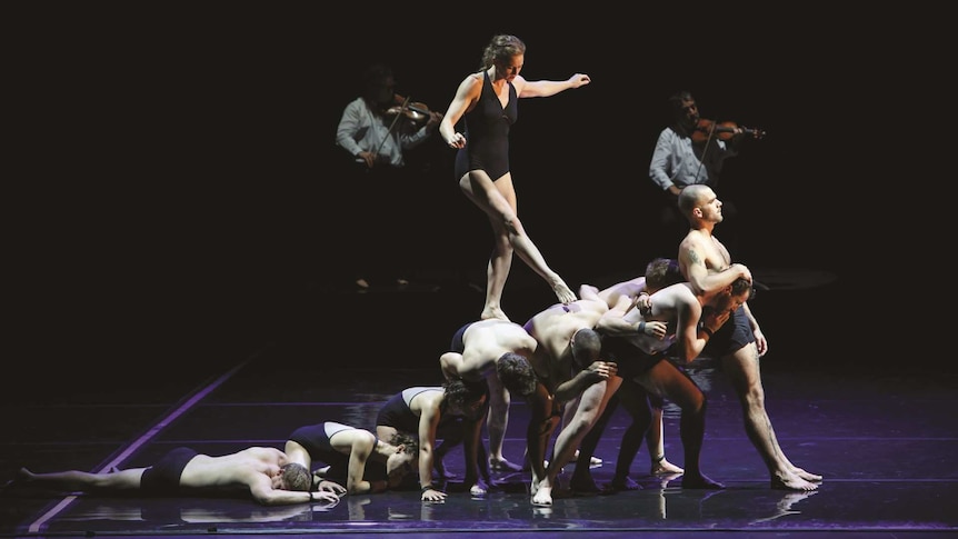 Eight circus performers use their bodies to form a ramp that another circus performer is walking up. Two violinists play behind.