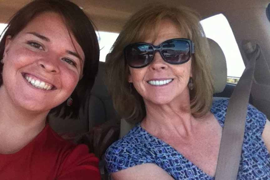 A woman with brown hair smiles in a car next to an older woman with dark blonde hair and sunglasses in a selfie.