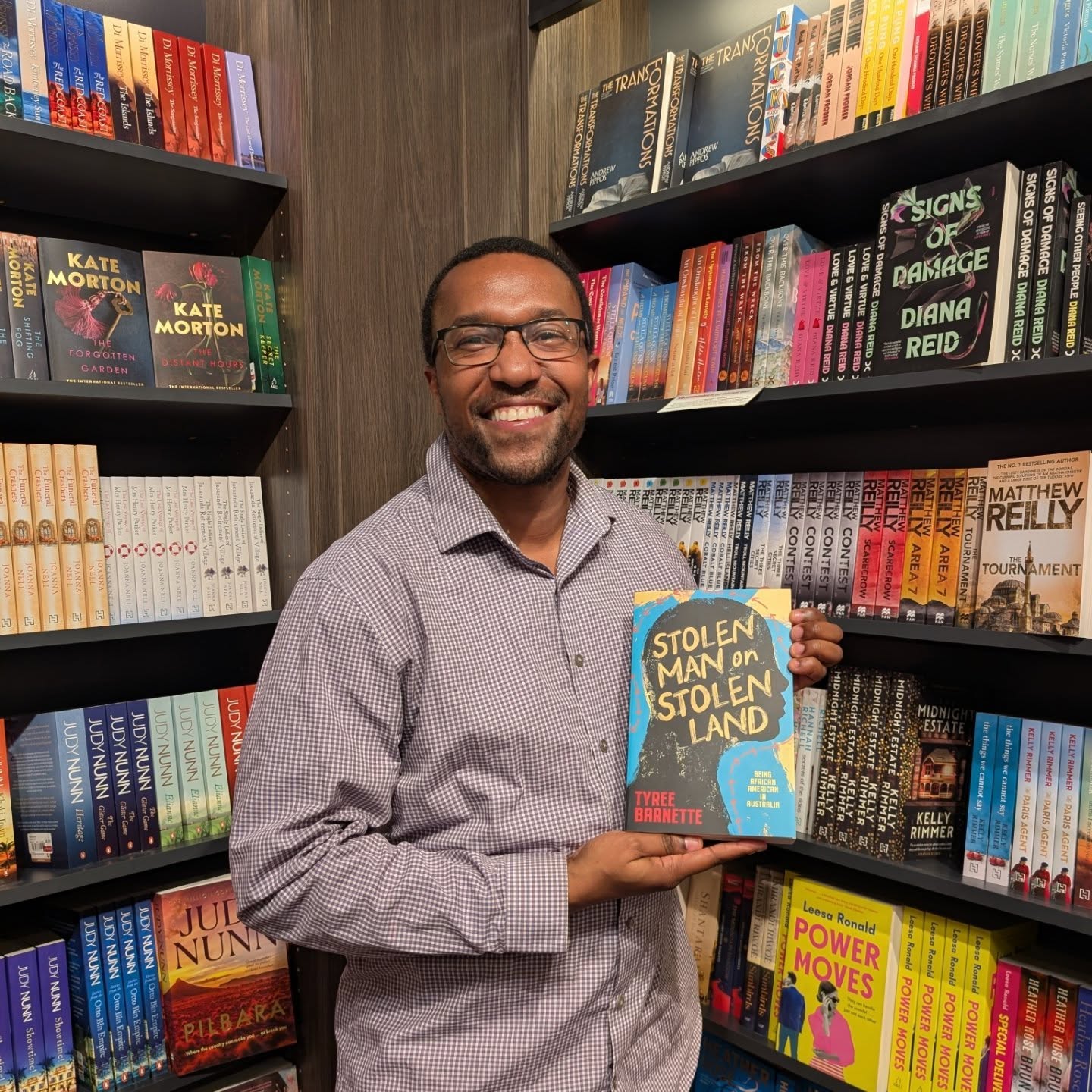 Tyree Barnette holding up his book in a bookstore, smiling, reading glasses
