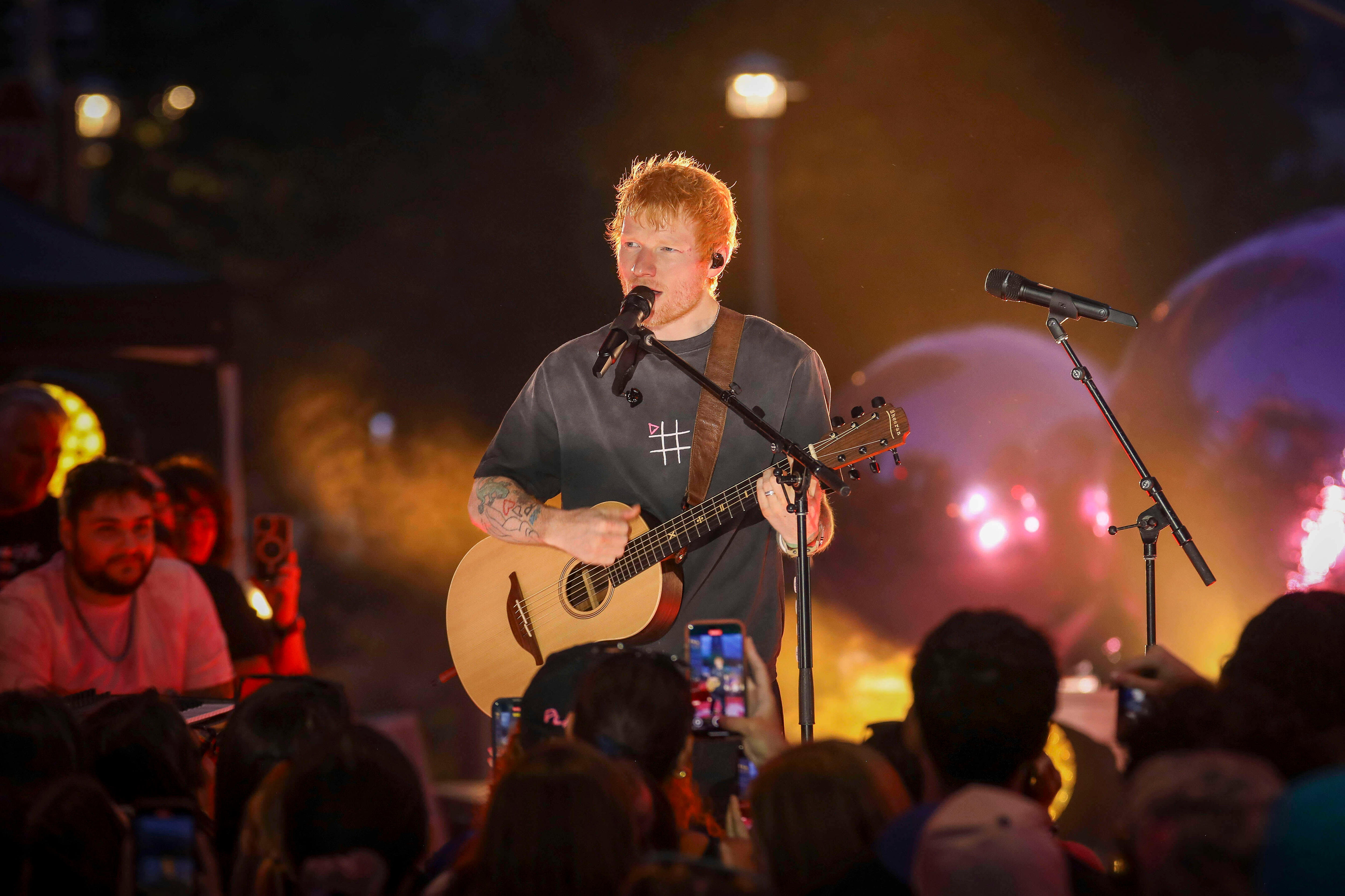 Ed Sheeran holds a guitar and sings into a microphone while on a stage.