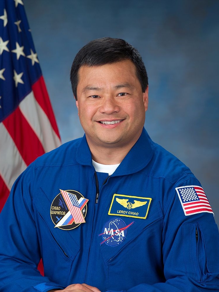 An Asian man poses for a photo in his NASA uniform, with the US flag on his right.