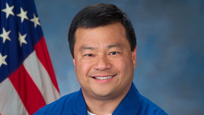 An Asian man poses for a photo in his NASA uniform, with the US flag on his right.