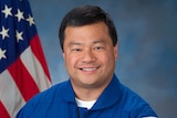 An Asian man poses for a photo in his NASA uniform, with the US flag on his right.