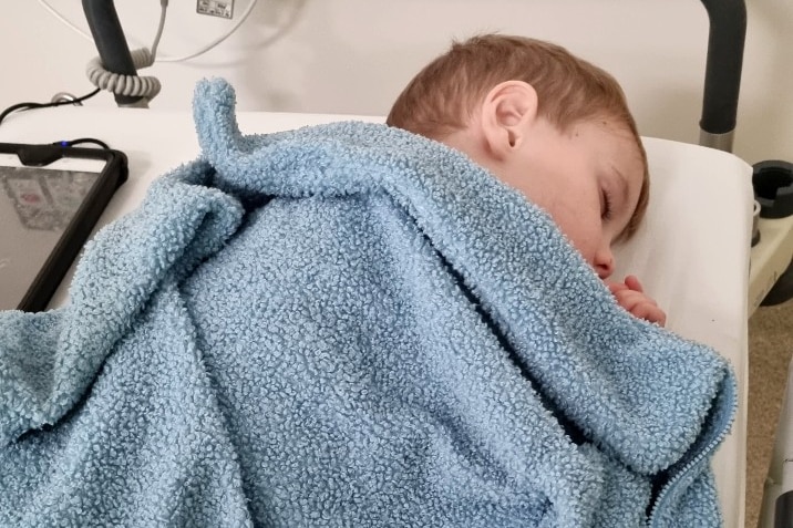 Teddy est allongé sur un lit d'hôpital, les yeux fermés.  Il est recouvert d'une couverture.