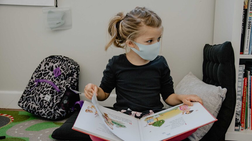 A child sits on the floor, reading a book, wearing a face mask.