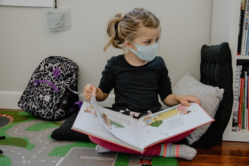 A child sits on the floor, reading a book, wearing a face mask.