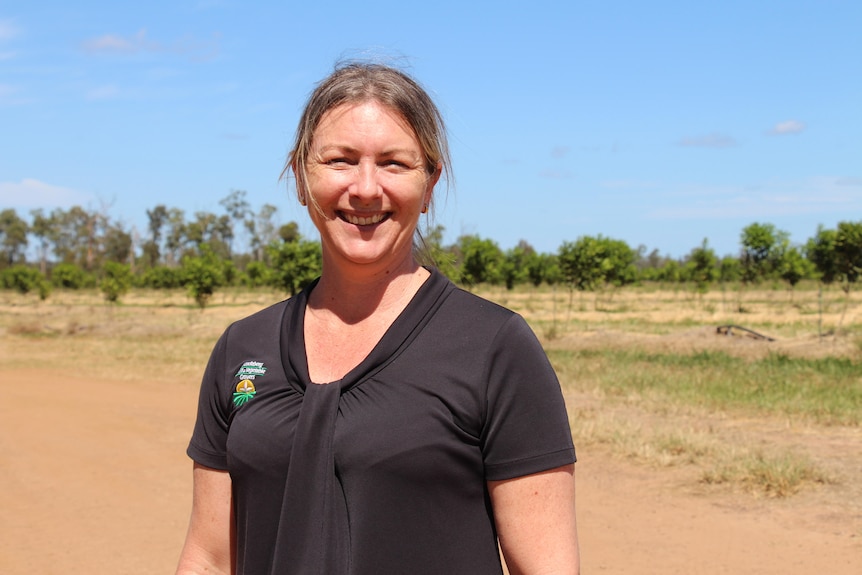A woman in a black shirt smiles at the camera, with red dirt and trees behind her.