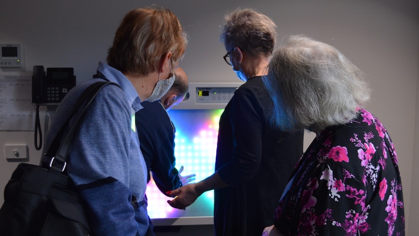 From behind, you view a group of three women watching on as an elderly man places his hand on a brightly-lit screen.