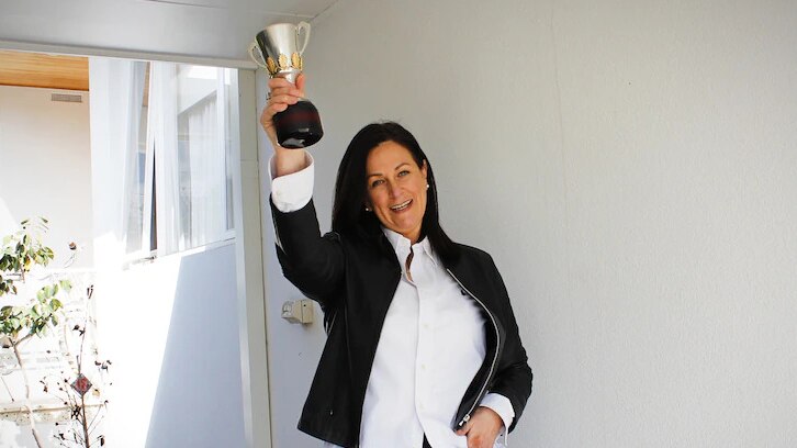 A woman with black hair holds a minuature version of the AFL premiership cup aloft. She is smiling triumphantly.