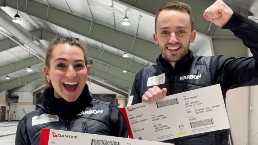 Tahli Gill and Dean Hewitt hold up oversized plane boarding passes while wearing puffy jackets in a curling arena.