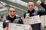 Tahli Gill and Dean Hewitt hold up oversized plane boarding passes while wearing puffy jackets in a curling arena.