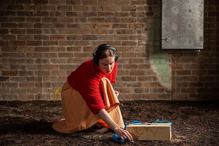 Colour photo of artist Megan Alice Clune kneeling down and interacting with device and soil in a dimly lit brick room.