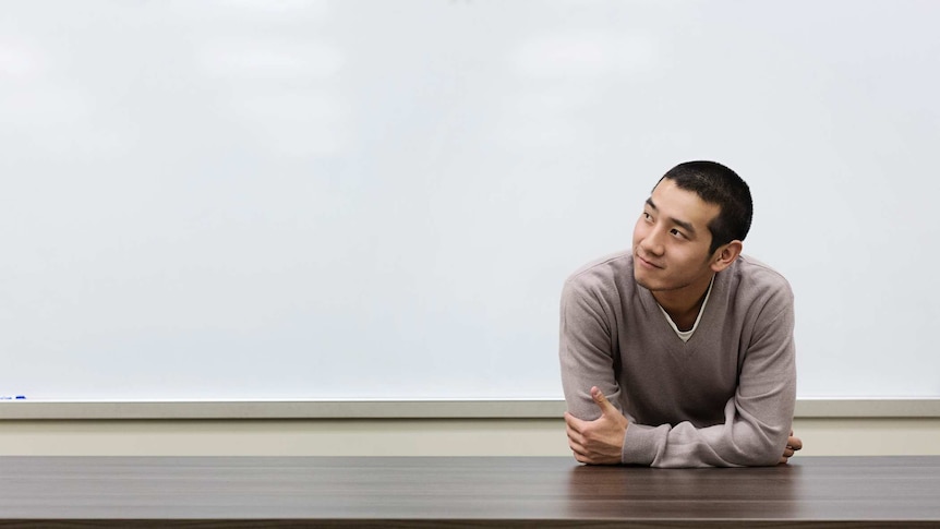 A young male Chinese student leans against a desk in a classroom and looks off into the distance.