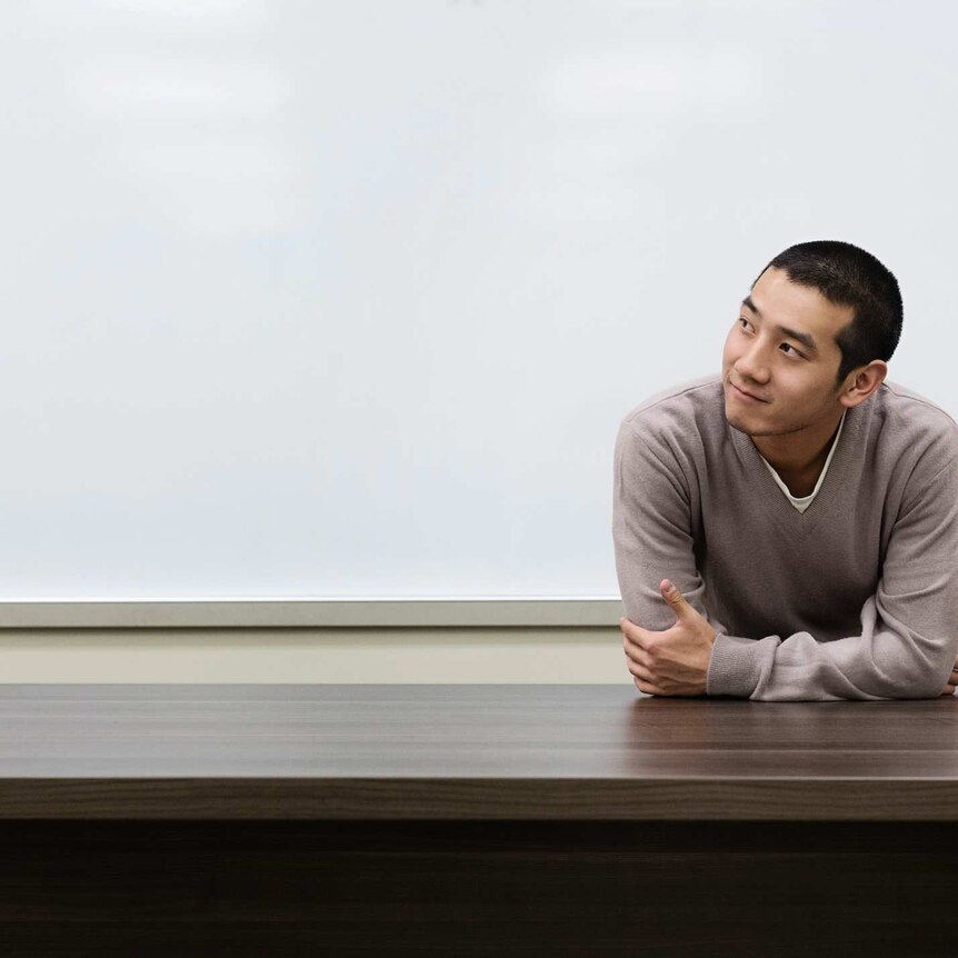 A young male Chinese student leans against a desk in a classroom and looks off into the distance.