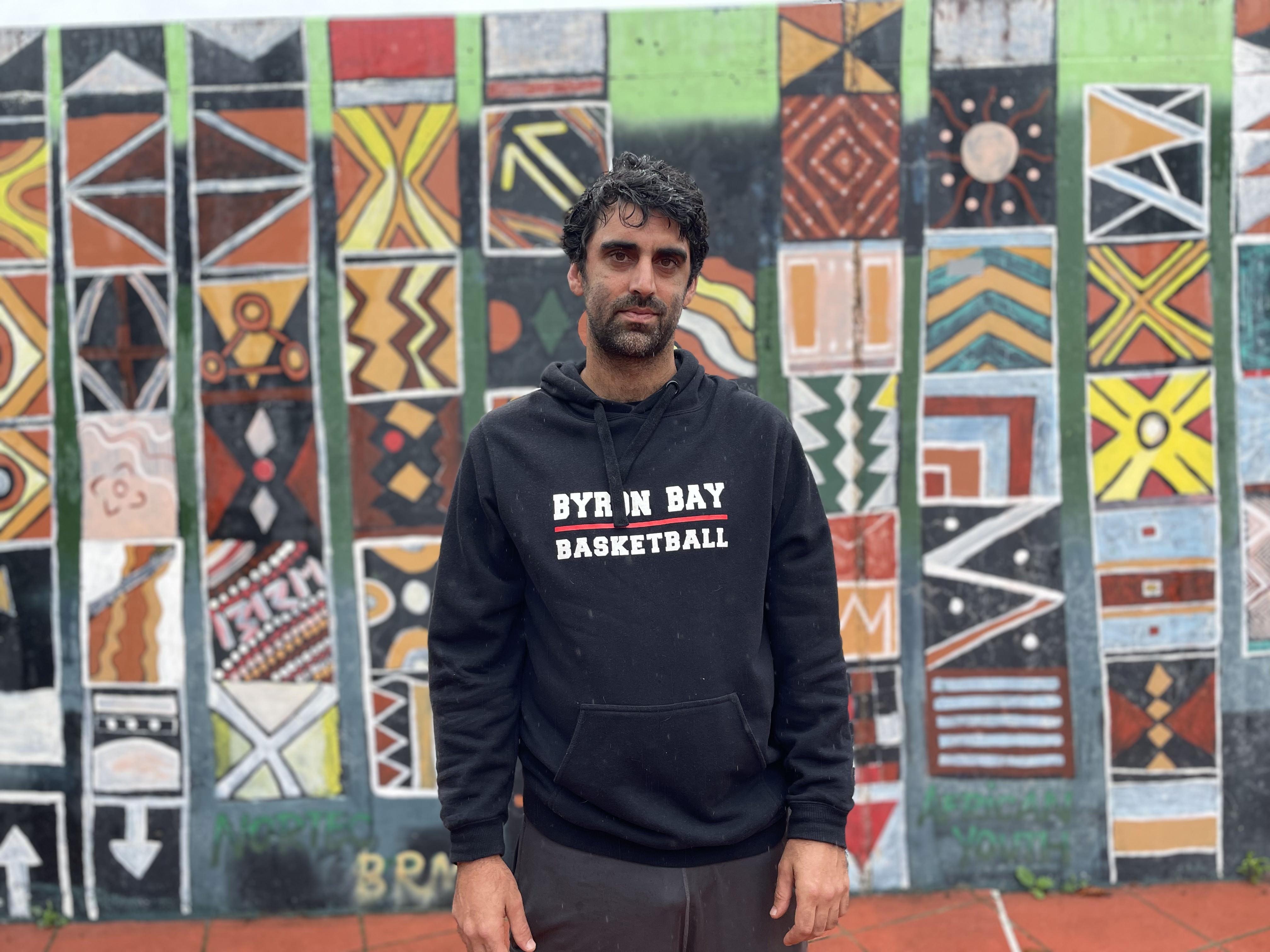 A photo of Surya standing in front of a tiled wall. He's wearing a black sweater that says 'Byron Bay Basketball'.