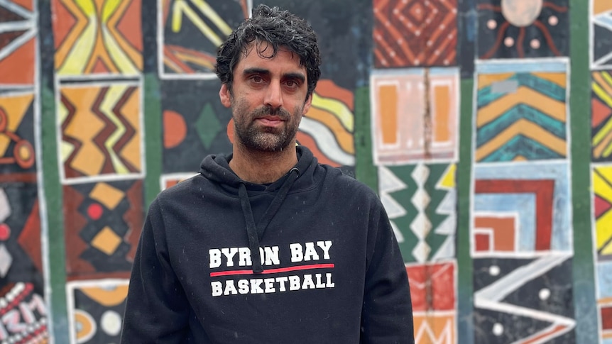 A photo of Surya standing in front of a tiled wall. He's wearing a black sweater that says 'Byron Bay Basketball'.