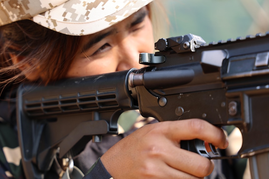 Woman in fatigues looks through scope in gun. 