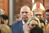 Dutton looks at a camera as he files into church among a crowd, a religious figure in traditional dress stands behind him.