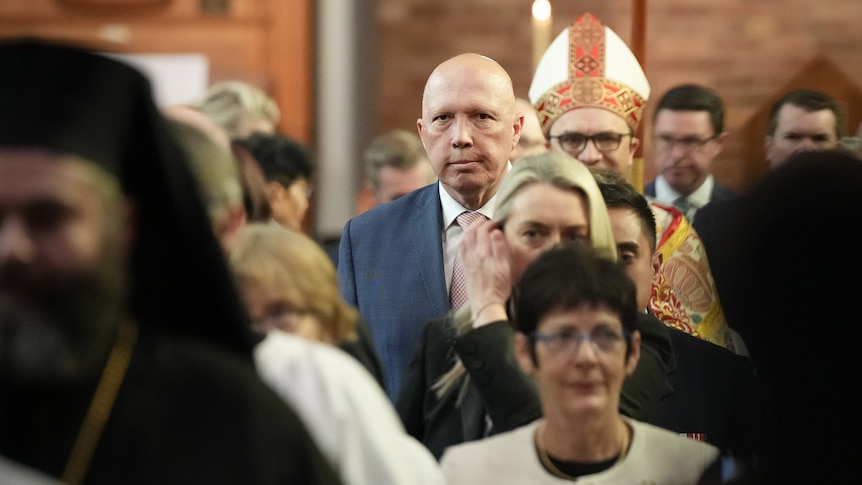 Dutton looks at a camera as he files into church among a crowd, a religious figure in traditional dress stands behind him.