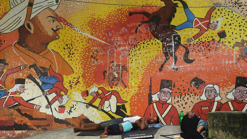 A family sleeps in front of a mural showing Tipu Sultan. It also shows British soldiers being injured.