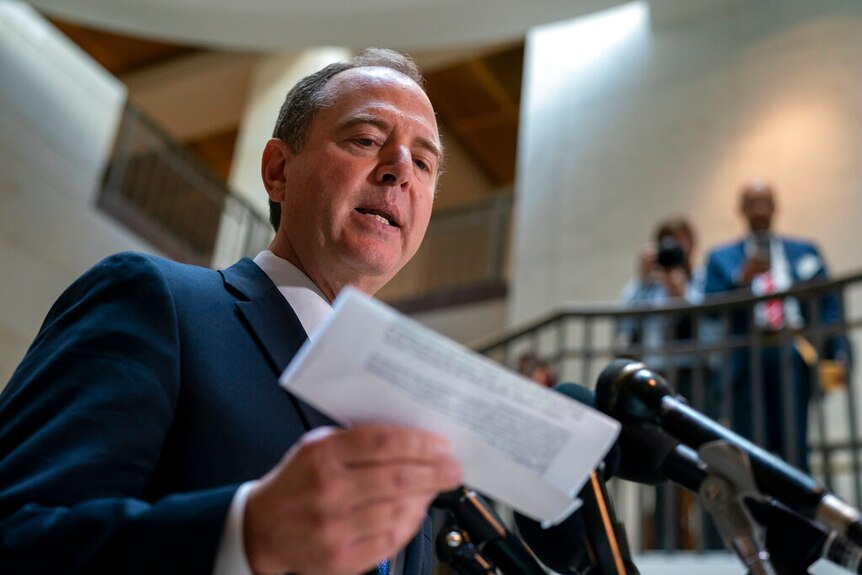 You look up at Adam Schiff as he speaks in front of a cluster of microphones while holding a folded white printed document.
