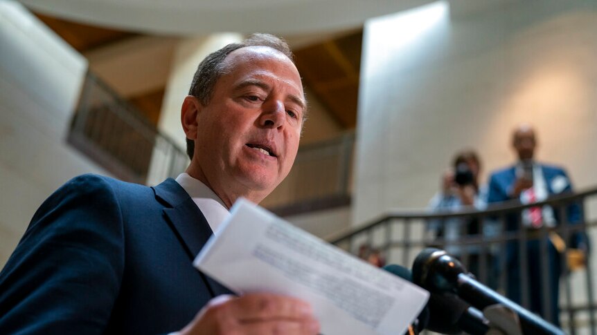Adam Schiff speaks in front of a cluster of microphones while holding a folded white printed document.