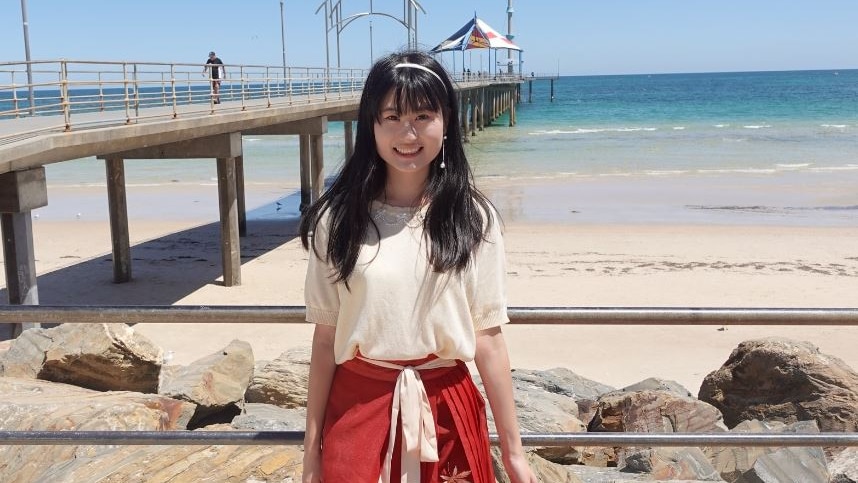 woman with long black hair, cream top and red skirt, posing in front of a jetty with beach in the background