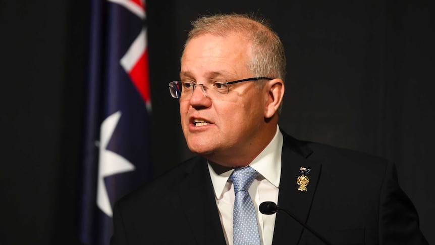 Scott Morrison looks to the left as he speaks in front of a dark backdrop with an Australian flag to the left.