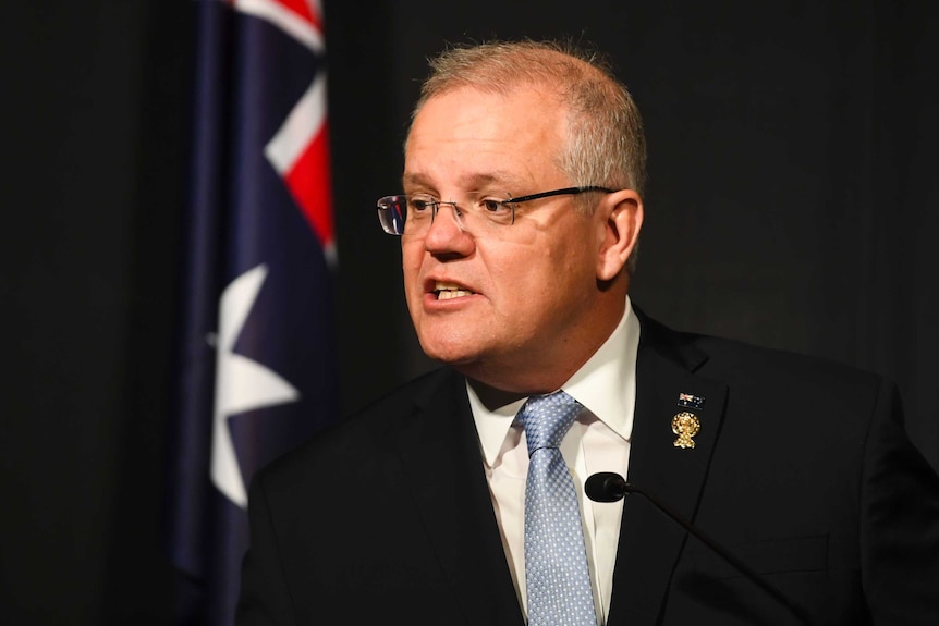 Scott Morrison looks to the left as he speaks in front of a dark backdrop with an Australian flag to the left.