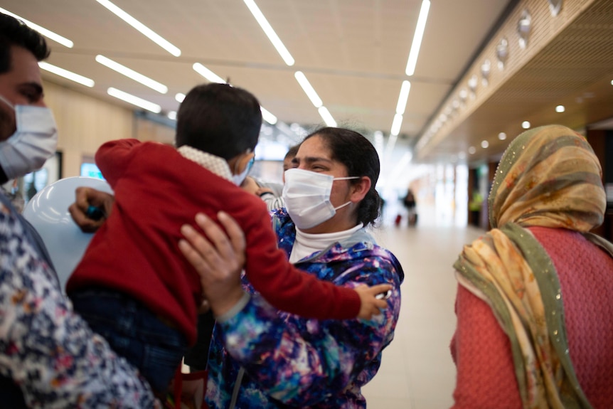 A masked woman at an airport reaches for a toddler for a hug