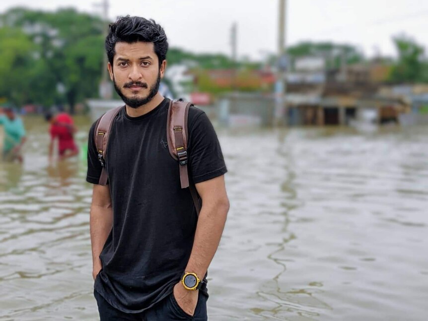 A young man in black T-shirt and jeans stands in a flooded street, looking straight to camera