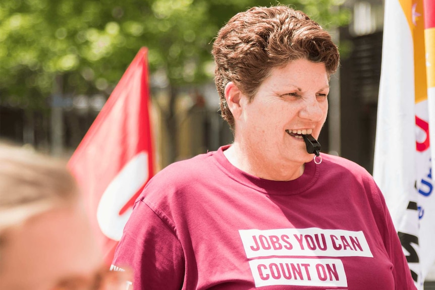 A woman with a a whistle in her mouth wears a tshirt with slogan 'jobs you can count on'. Blurred flags and trees in background.