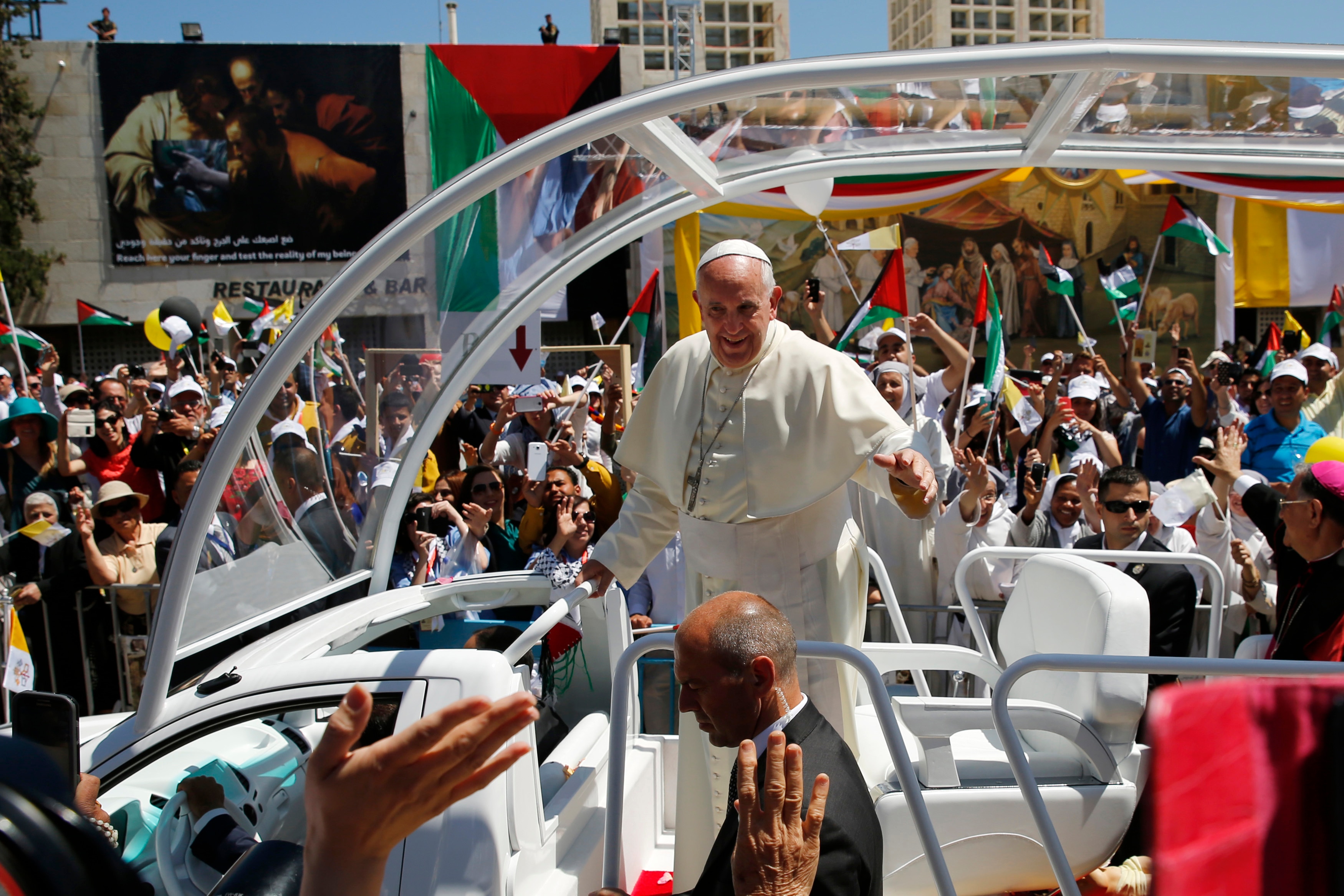 Pope Francis standing in the popemobile which has open sides and a glass roof surrounded by crowds with palestinian flags