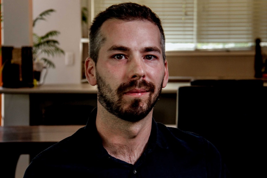 Gilles Fischer, bearded with black hair and wearing a black shirt, sits in lounge room with table and window with blinds visible