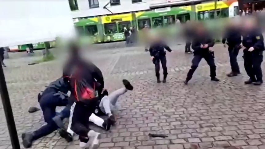 A man stands over two men falling to the ground, as police offers watch, in a paved square. Everyone's faces are blurred.