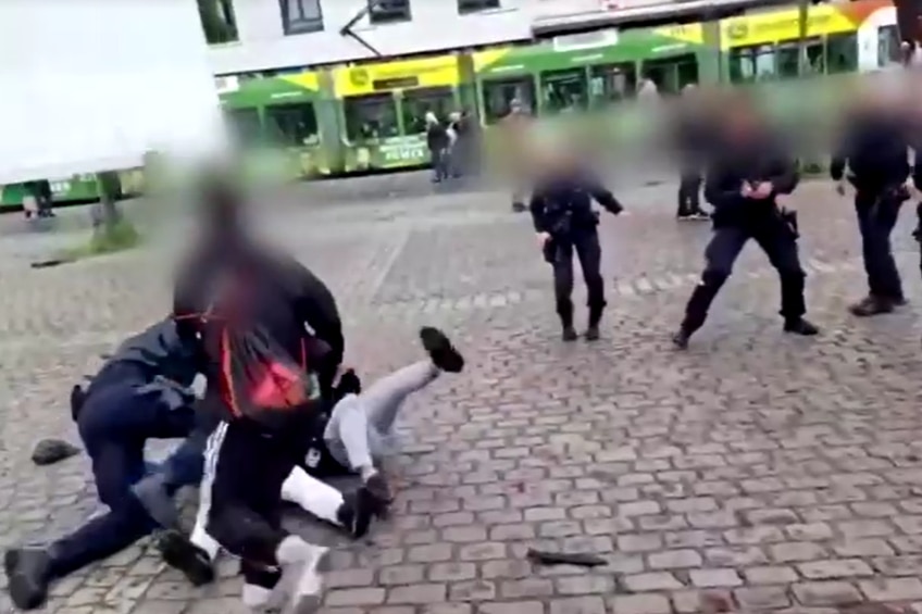 A man stands over two men falling to the ground, as police offers watch, in a paved square. Everyone's faces are blurred.