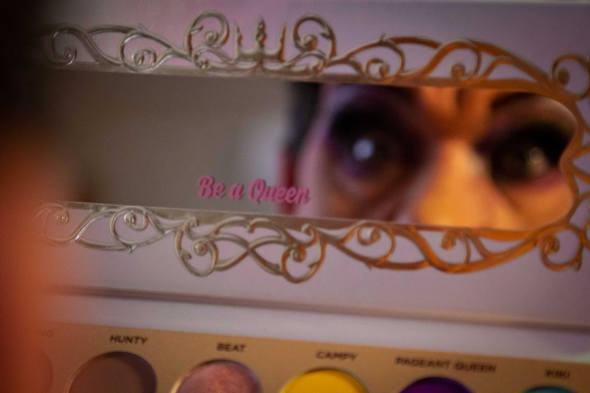 Drag queen MadB is looking into a makeup mirror.