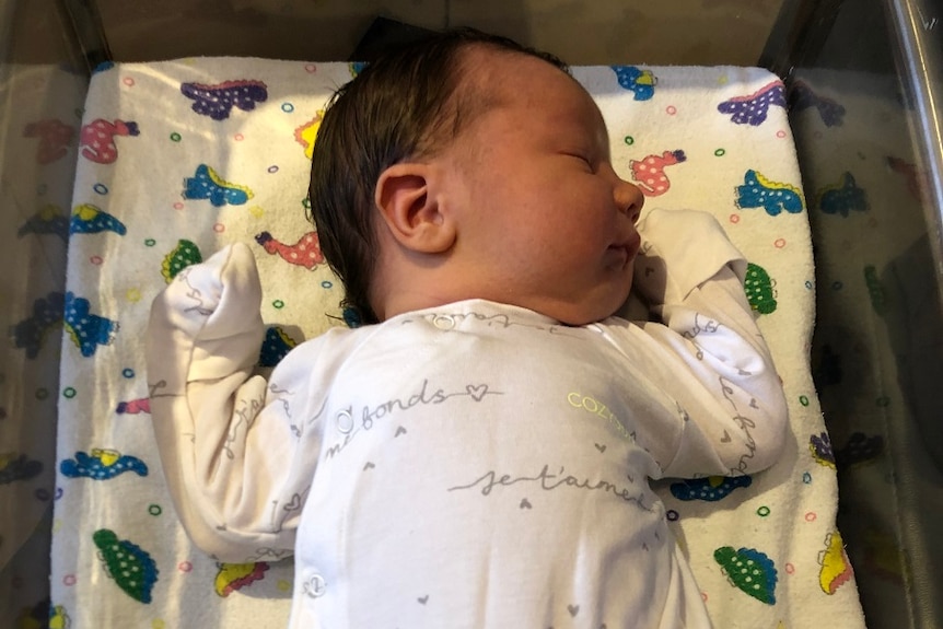 Newborn baby with brown hair wearing white one piece lies asleep iin a hospital cot with a colourful sheet.