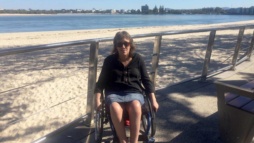 A woman in a wheelchair sits on a boardwalk with a sunny white sand beach, clear blue water and skies and buildings behind her.